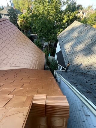 Aerial close-up of Interlock Copper Penny aluminum slate roofing showing a complex hip transition and copper ridge cap detail beside a neighboring asphalt shingle roof.