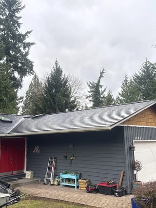 Black Interlock Slate aluminum roof being installed on a grey single-storey home with a metal roof among tall trees, Redmond, Washington
