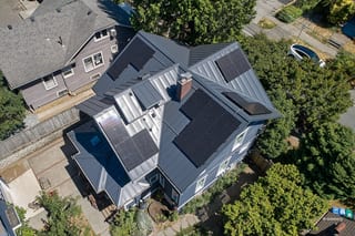 Deep Charcoal Interlock Standing Seam aluminum roof on a grey two-storey home with dormers among tall trees in, Seattle, Washington