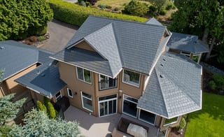 Deep Charcoal Interlock Slate aluminum roof on a tan two-storey home with dormers among tall trees, Blaine, Washington