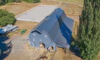 Aerial view of a large gambrel-style agricultural barn with black Interlock slate metal roofing and an adjacent riding arena in rural Longview, WA.