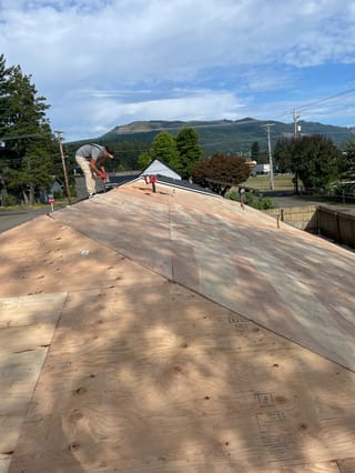 Roofer installing new plywood decking on a steep gable roof with mountain backdrop in Morton, WA, prior to Interlock Copper Penny aluminum slate application.