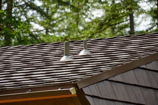 Close-up of two metal vent pipe flashings on a Colonial Brown Interlock aluminum shake roof with lush forested backdrop in Yacolt, WA.