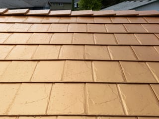 Close-up of Interlock aluminum slate metal roofing in Copper Penny finish, showing rain-beaded tiles with ridge cap detail against a suburban background.