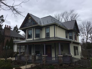 Weathered Interlock Slate aluminum roof on a cream two-storey home with dormers and a porch among tall, Mayville, Wisconsin