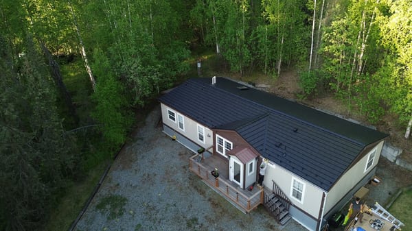 Metal roof on a ranch-style house in a forested rural area with lush greenery and a blue sky.