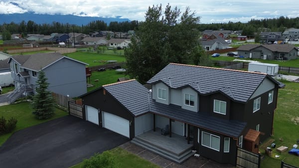 A modern single-family home with a metal roof in a suburban area surrounded by lush greenery and a blue sky.
