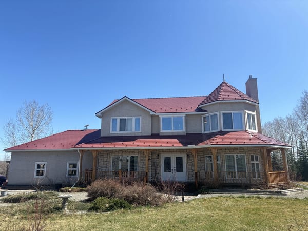 Two-storey rural acreage home with complex Tile Red Interlock aluminum slate metal roof featuring turret, dormers, and wraparound porch in Foothills County, AB.