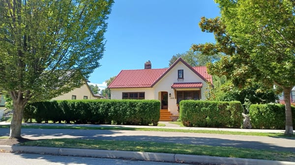 1.5-storey stucco cottage with Interlock Tile Red Mediterranean aluminum tile roof, chimney, and arched entry, framed by mature trees in Kelowna, BC.