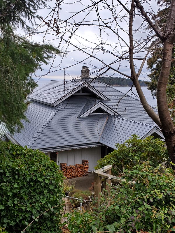 Deep charcoal Interlock aluminum slate metal roof on a complex multi-hip craftsman home in Sechelt BC, with chimney, dormers, and ocean backdrop.