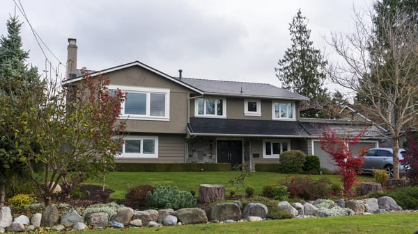 Two-storey suburban home in Tsawwassen BC with black Interlock aluminum shingle metal roof, stucco exterior, chimney, and landscaped front yard.