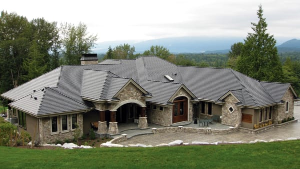 Large single-story stone estate in Mission BC with complex multi-hip charcoal grey Interlock aluminum slate metal roof, chimney, and mountain backdrop.