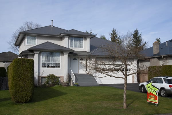 Two-storey colonial-style suburban home with Interlock Musket Grey aluminum slate multi-hip roof, white stucco exterior, and Interlock yard sign in Surrey, BC.