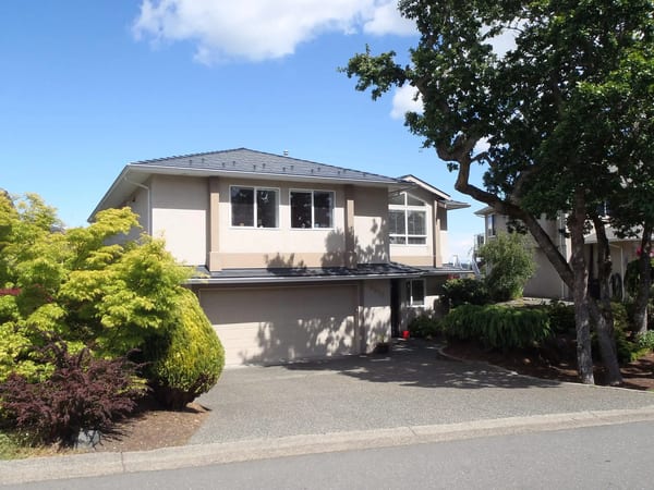Two-storey suburban home in Victoria BC with a multi-hip deep charcoal Interlock aluminum slate metal roof, beige stucco exterior, and mature landscaping.