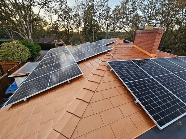 Aerial view of a Copper Penny Interlock aluminum slate metal roof with multiple solar panels and a red brick chimney on a suburban home surrounded by trees.