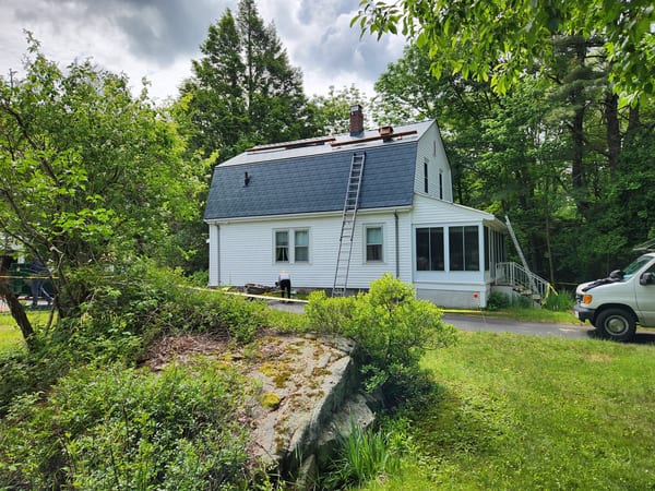 Hunter green Interlock aluminum slate metal roof being installed on a white Cape Cod home with gambrel profile and brick chimney, surrounded by trees in Sharon, MA.