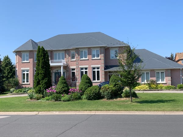 Large two-storey brick Colonial home in Markham ON with Interlock deep charcoal aluminum slate multi-hip roof, manicured landscaping, and balcony over entry.