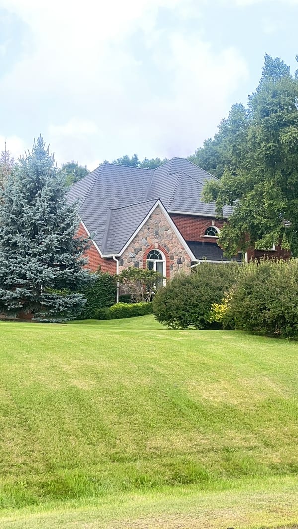 Black Interlock aluminum slate metal roof on a 1.5-storey Tudor-style brick-and-stone home surrounded by mature trees in Campbellville, ON.
