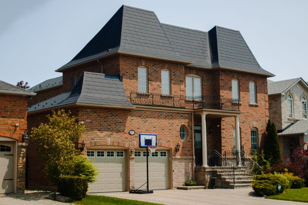 Two-storey brick Colonial home in Woodbridge ON with a complex multi-hip black Interlock aluminum slate metal roof and prominent Mansard-style lower sections.