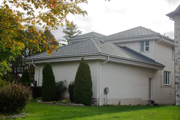 Rear-left view of a two-storey white brick home in Whitby, ON featuring Interlock charcoal grey aluminum shake roofing across a complex multi-hip roof.