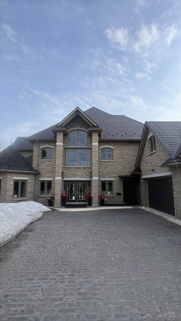 Two-storey Tudor-style stone home in Sudbury ON with complex multi-hip Interlock Tudor Brown aluminum slate roof and cobblestone driveway in winter.