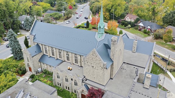 Aerial view of a Gothic stone church in Toronto with a newly installed Interlock deep charcoal aluminum slate metal roof, green copper steeple, and complex multi-plane roofline.