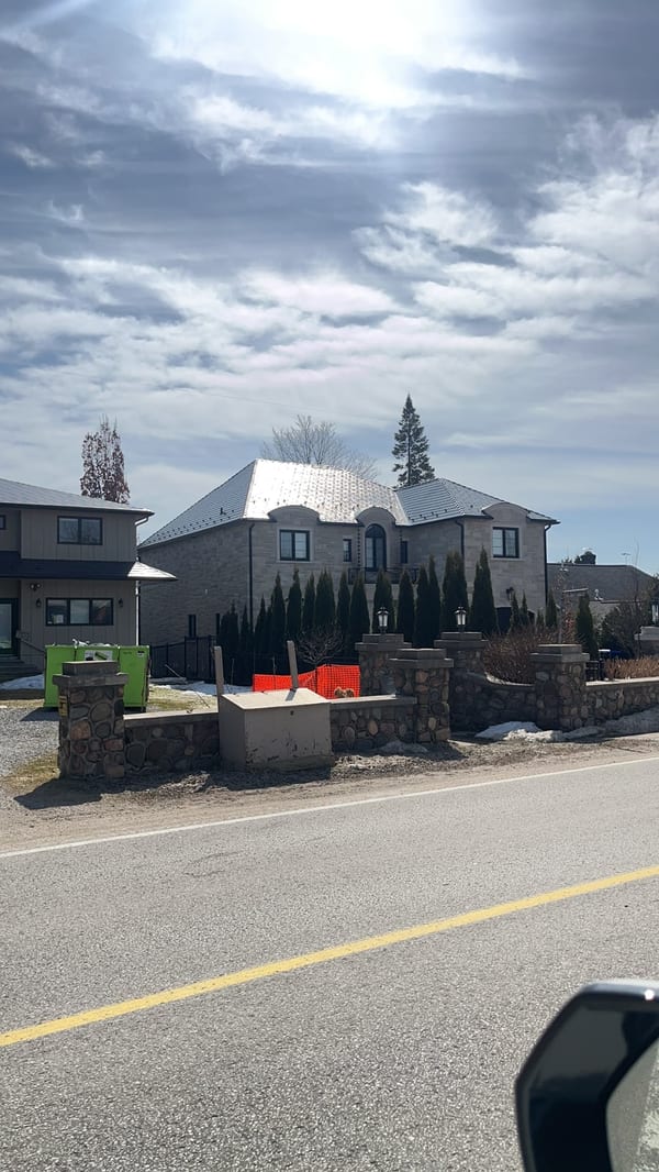 Street-view of a large two-storey stone Mediterranean home in Innisfil, ON with a steeply pitched multi-hip Deep Charcoal Interlock aluminum cedar shingle metal roof.