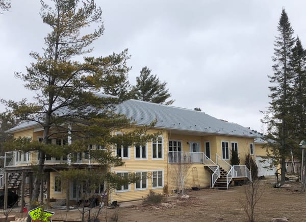 Two-storey yellow cottage with Interlock deep charcoal cedar shingle aluminum hip roof, white railings, and large decks near Wasaga Beach.