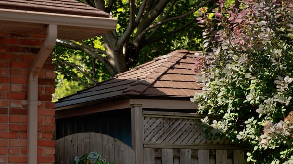 Interlock Colonial Brown cedar shingle aluminum hip roof on a small gazebo or hot tub enclosure beside a brick home, surrounded by lush garden greenery.