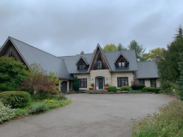Front elevation of a Tudor-style stone home in St. George, ON featuring a steeply pitched Interlock aluminum slate roof in charcoal black with multiple dormers and complex valleys.