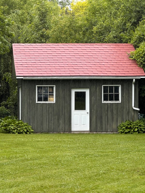 Front elevation of a rustic wood-clad outbuilding with a Tile Red Interlock aluminum shingle gable roof, white door, and twin windows in Almonte, ON.