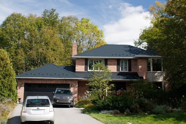 Two-storey brick Colonial home in Kingston, ON with a black Interlock aluminum slate metal roof, chimney, and lush tree-lined suburban setting.