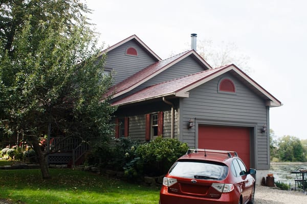 1.5-storey grey craftsman home in Kingston ON with Tile Red Interlock aluminum slate metal roofing, matching garage door, and lakeside backdrop.
