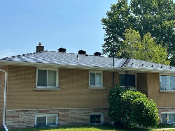 Black Interlock aluminum slate metal roof on a single-storey ranch-style brick home in Cornwall, ON, featuring multiple roof louvers and a brick chimney.