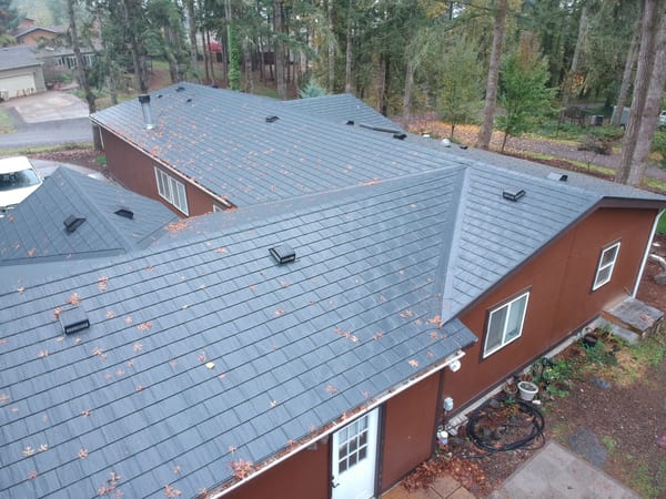 Aerial view of a complex multi-hip Interlock aluminum slate roof in charcoal black on a red-sided home surrounded by tall firs in Albany, OR.