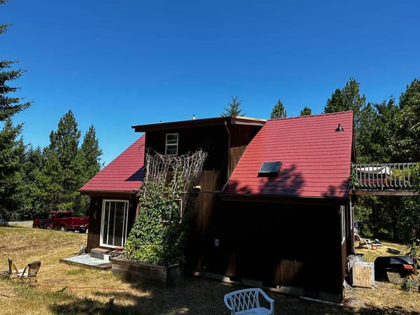 Two-story chalet-style home in Eugene, OR with steep Tile Red Interlock aluminum slate roof, dormer, and skylight surrounded by tall evergreens.