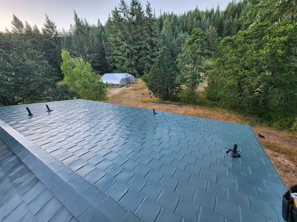 Aerial view of a hunter green Interlock aluminum slate metal roof with three vent pipes, surrounded by dense Pacific Northwest forest in Eugene, OR.