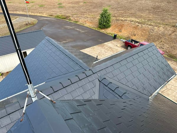 Aerial drone view of a complex multi-hip black Interlock aluminum slate metal roof with multiple valleys, snow guards, and ridge caps on a rural single-family home.