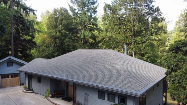 Aerial front view of a ranch-style home with Interlock Musket Grey aluminum slate metal roofing, hip roof, and wooded backdrop in Eugene, OR.