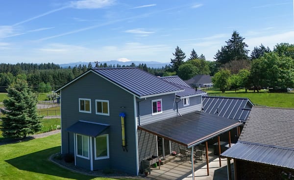 Black Interlock Standing Seam aluminum roof on a grey two-storey home with dormers among tall pines, Warren, Oregon