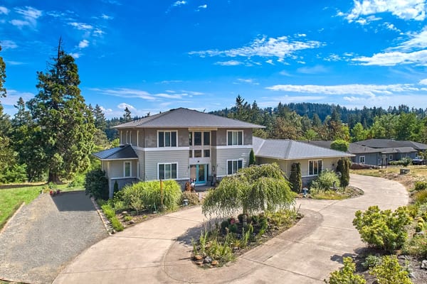Two-story Oregon City home with multi-hip black Interlock aluminum slate metal roof, circular concrete driveway, and lush landscaping backed by evergreen forest.