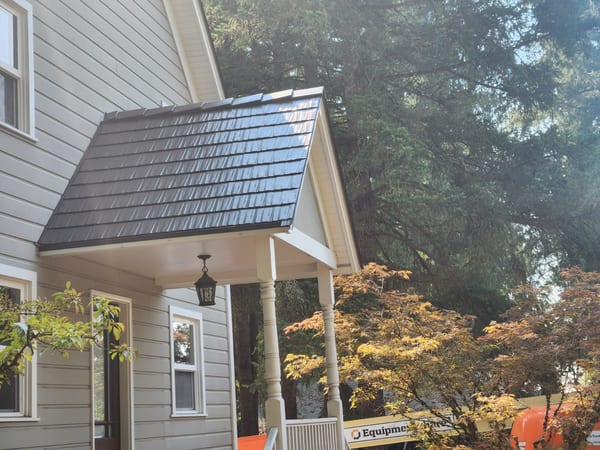 Interlock Tudor Brown cedar shingle metal roof on a 2-story Craftsman home with a hip-roofed front porch entry, surrounded by tall conifers in North Plains, OR.