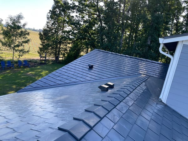 Aerial view of a deep charcoal Interlock aluminum slate metal roof with complex hip valleys on a rural Craftsman home surrounded by vineyard and trees.
