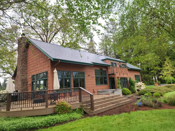 Craftsman home in Corvallis OR with Interlock deep charcoal aluminum slate roof, stone chimney, large rear deck, and hot tub amid lush forest.