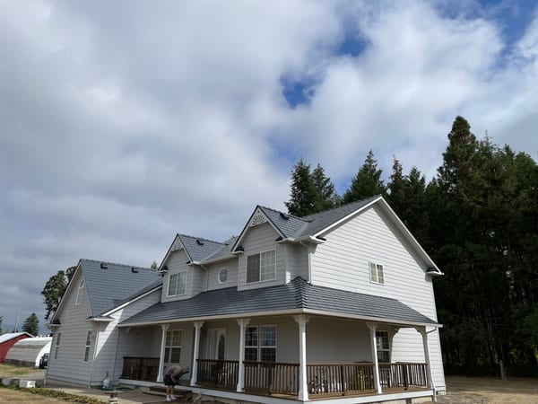 Two-story Craftsman farmhouse with a complex deep charcoal Interlock aluminum slate metal roof featuring multiple gables, dormers, and a covered front porch, set among tall evergreens in rural Oregon.