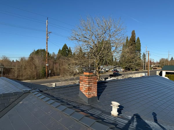 Deep charcoal Interlock aluminum slate metal roof with brick chimney flashing detail, skylights, and vent pipe on a suburban Beaverton home.