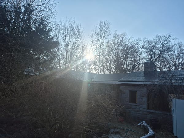 Deep charcoal Interlock aluminum cedar shingle metal roof on a 1-story stone heritage home in Harrisville RI, with chimney, surrounded by bare winter trees.