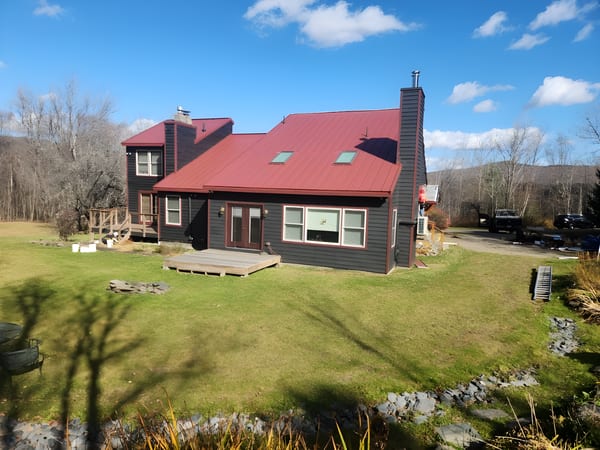 Rear elevation of a dark-sided Vermont chalet with a steep tile-red Interlock aluminum standing seam roof, twin chimneys, and skylights.