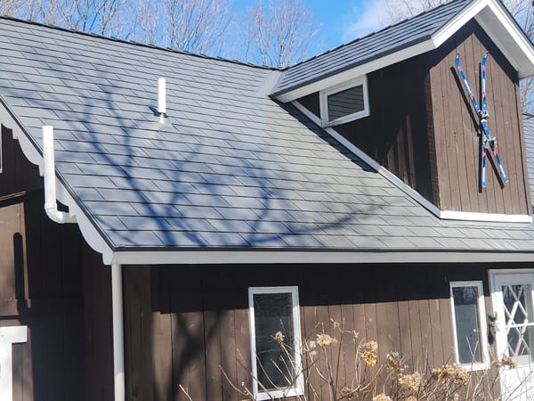 Dark brown chalet-style Vermont home with Interlock Musket Grey aluminum cedar shingle metal roof, white gutters, valley detail, and decorative skis on siding.