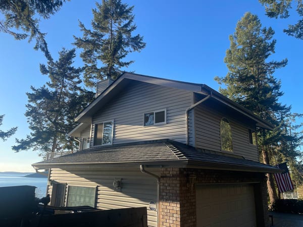 Two-story waterfront home in Anacortes WA with Interlock deep charcoal aluminum slate metal roofing, surrounded by tall evergreens with Puget Sound views.